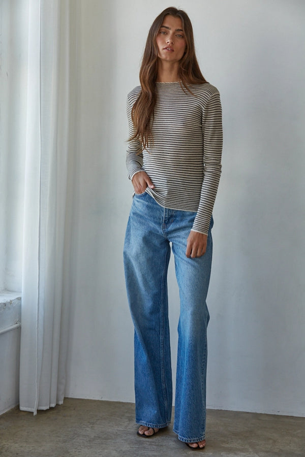 Woman wearing a striped sweater and blue jeans standing in a room with white walls and a window.