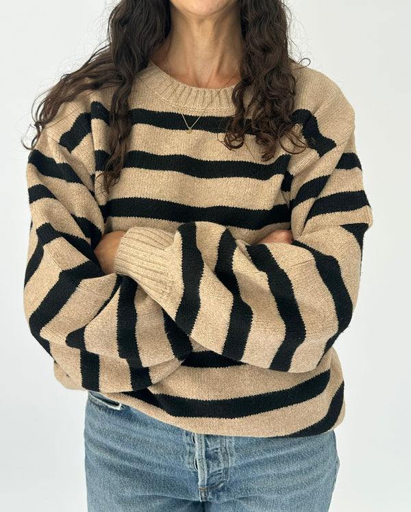 A woman wearing a beige knit sweater with bold black horizontal stripes, paired with blue jeans, shown from the neck down against a plain white background.