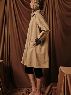 A woman stands in profile wearing a mid-length beige trench coat over black cropped pants, posed against draped brown fabric with studio props at the sides.