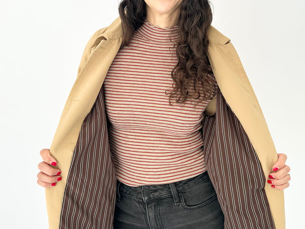 A woman wearing a beige trench coat over a red and taupe striped mock neck top, holding the coat open against a plain white background.